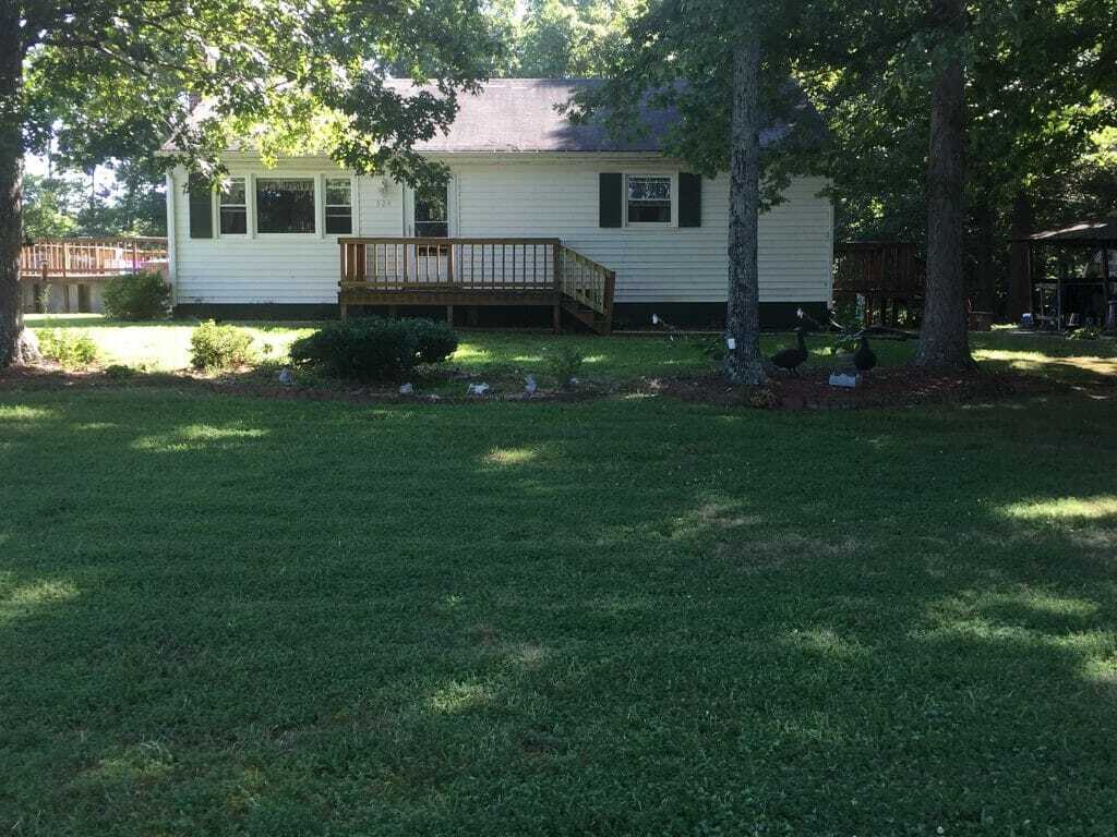 A small white house with a front porch sits behind a wide, well-kept lawn, shaded by several large trees belonging to Sponsored Residential Providers Beth and Otis Fowler in Rustburg, Virginia.