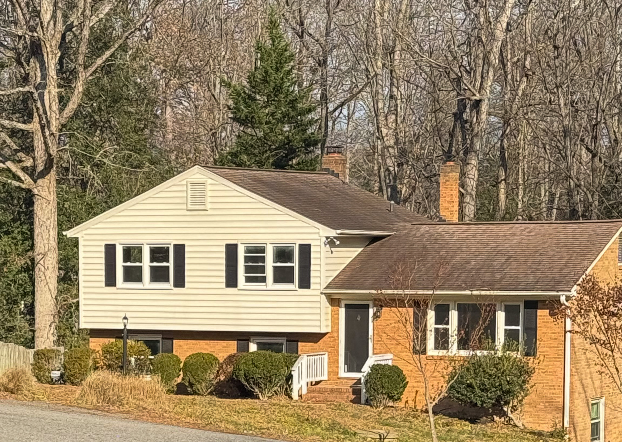 A split-level house with tan siding, red brick, black shutters, a dark roof, and trimmed shrubs, set against tall leafless trees belonging to Sponsored Residential Provider Selena Banks in Chesterfield, Virginia.