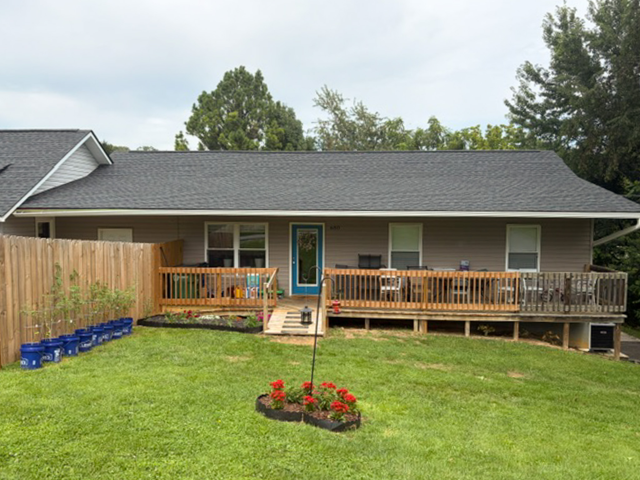 Single-story home with gray siding and a dark shingled roof. A wooden deck with ramp access spans the front of the house, overlooking a large green lawn with landscaped flower beds and a wooden privacy fence belonging to Group Home Provider Luke Haynes in Christiansburg, VA.