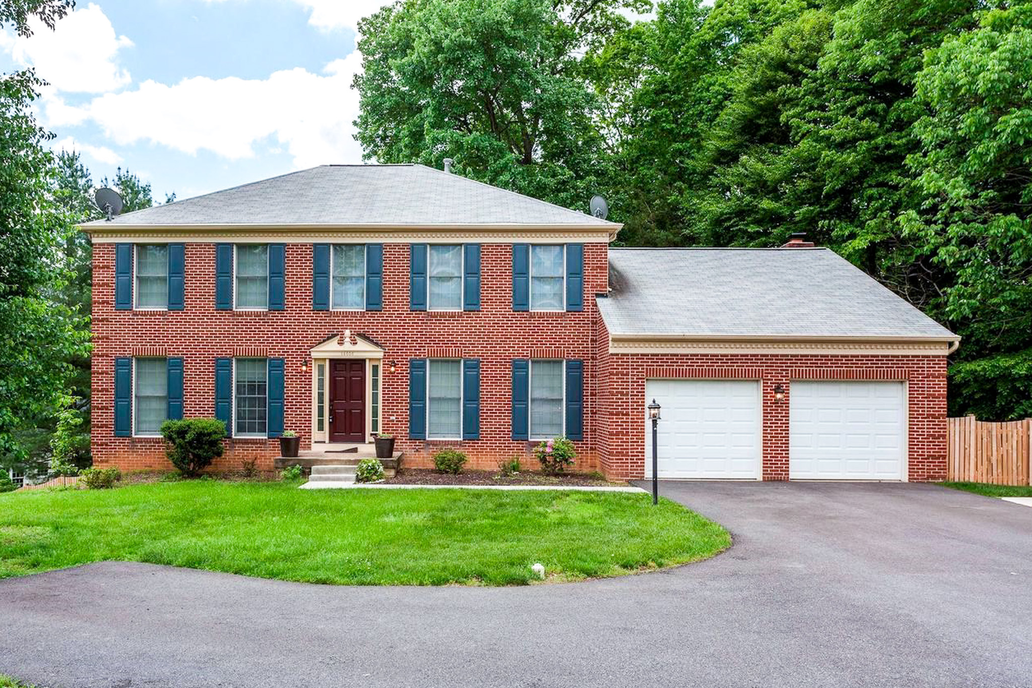 Two-story red brick house with blue shutters, a two-car garage, and a green lawn surrounded by trees inside the home of Sponsored Residential Provider Gamu Gomo in Woodbridge, Virginia.