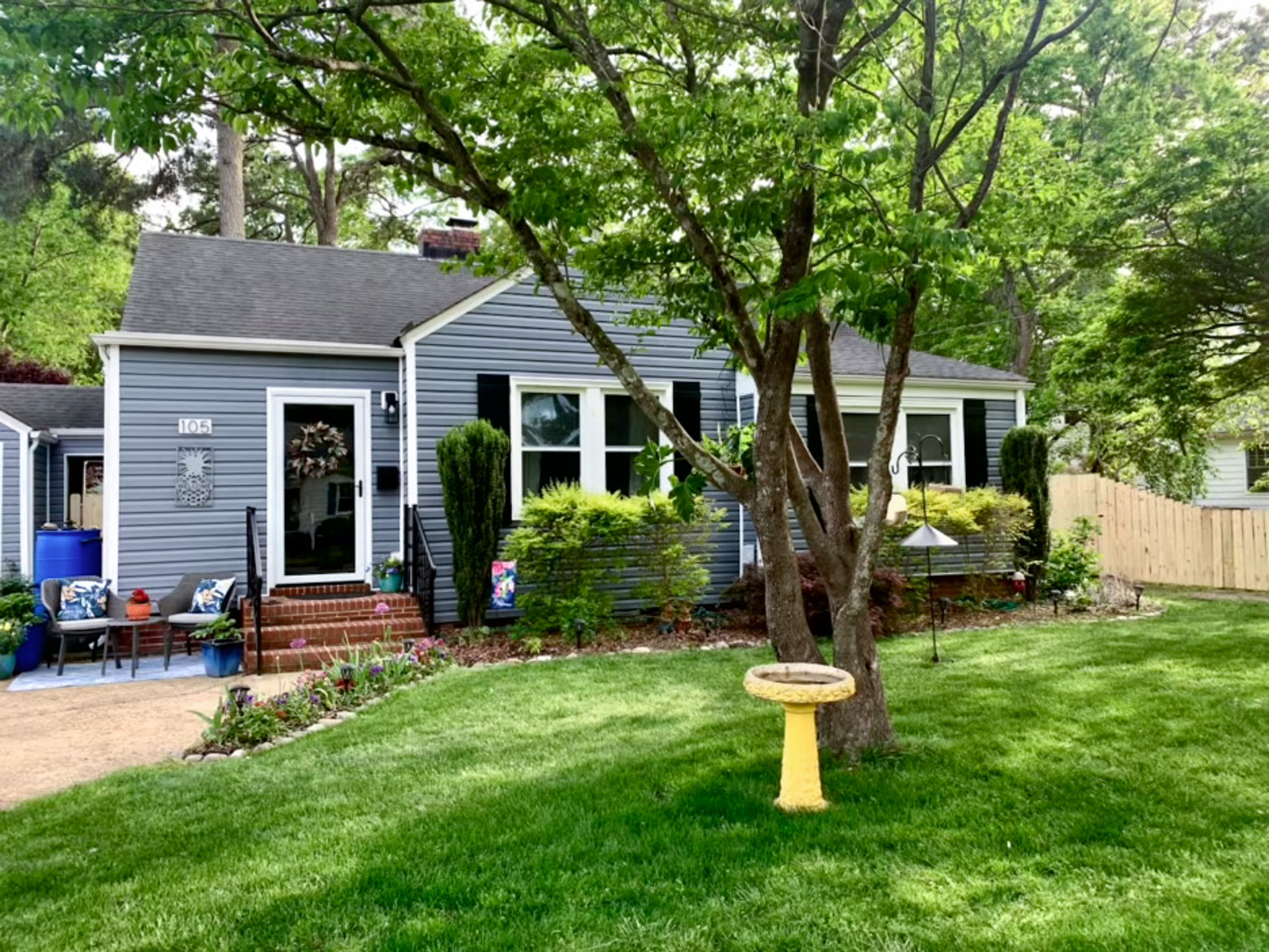 Gray single-story house with white trim, shaded by trees, featuring a green lawn, birdbath, and small front patio belonging to Sponsored Residential Provider Venus Wilding in Norfolk, Virginia.
