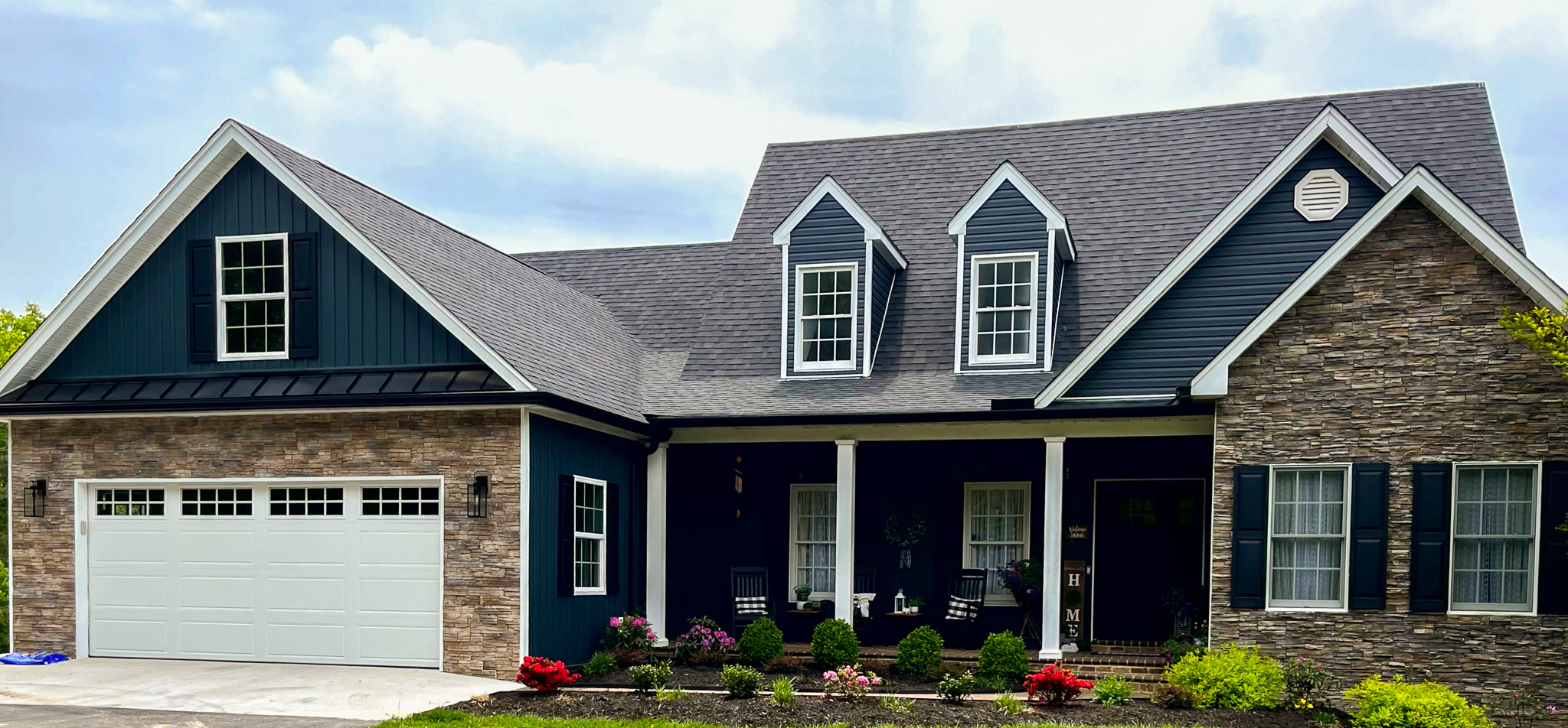 Modern house with dark blue siding, stone accents, dormer windows, and a covered front porch beside a white two‑car garage belonging to Group Home Providers Sheila and Steve Wagner in Evington, Virginia.