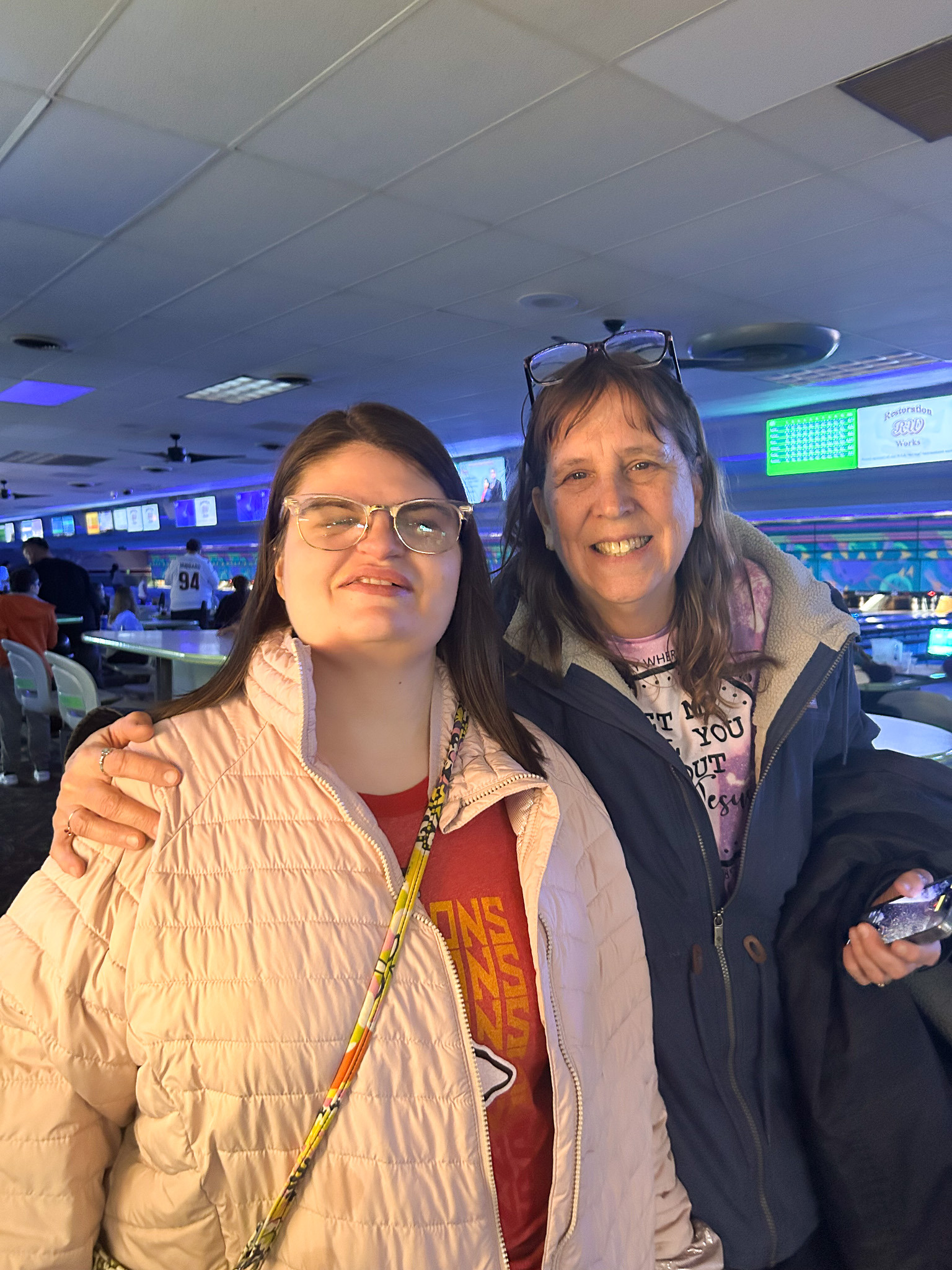 Two womenstanding together inside a bowling alley, wearing winter jackets, with bowling lanes and score screens lit up in the background.