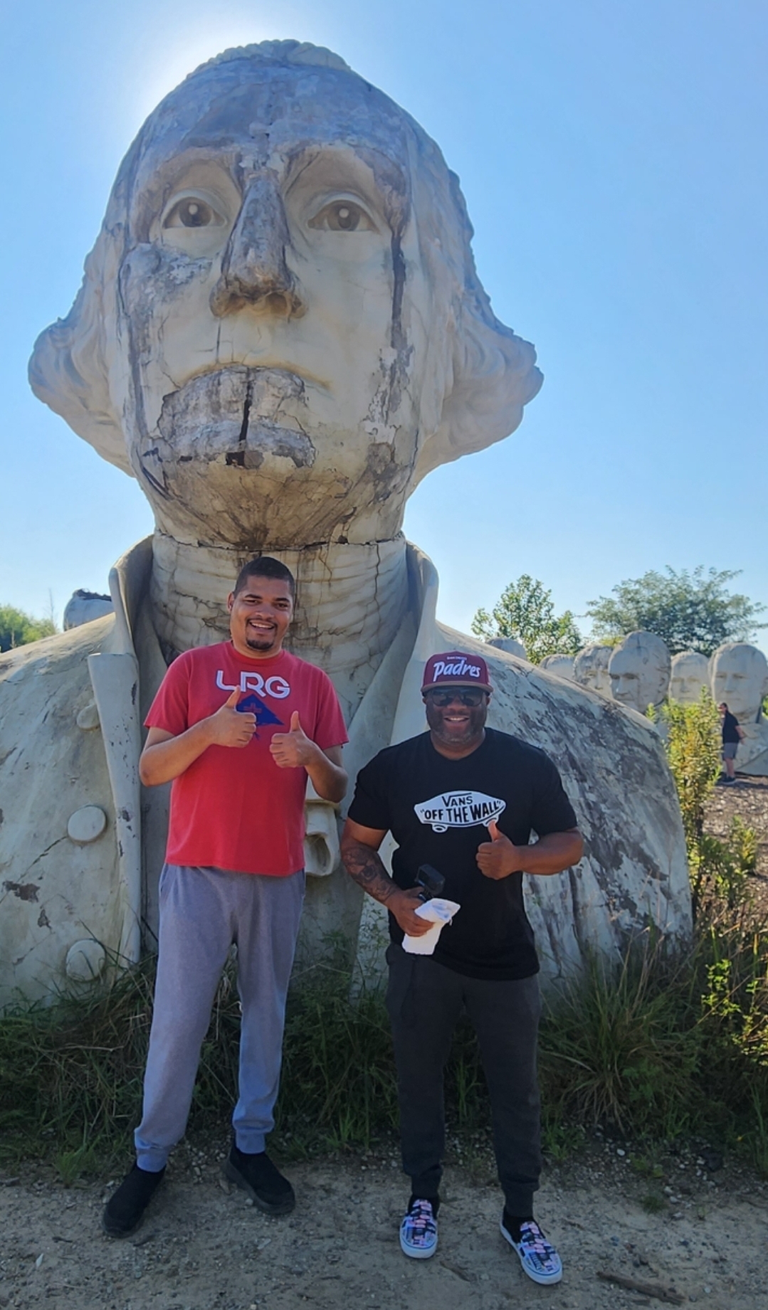 One man in a red shirt and one man in a black shirt and red hat giving a thumbs up while smiling in front of a statue outdoors