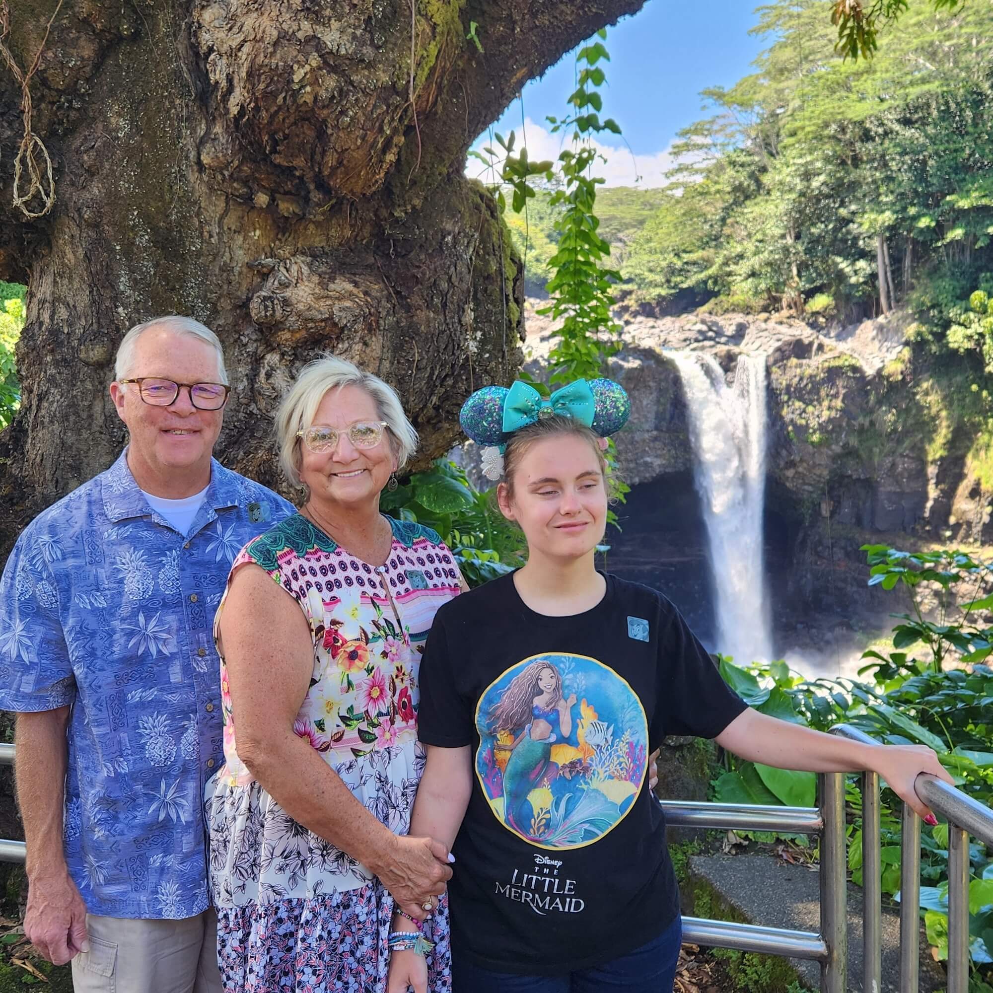 A mom, dad, and adult daughter stand in front of a waterfall and tree at Disney World.