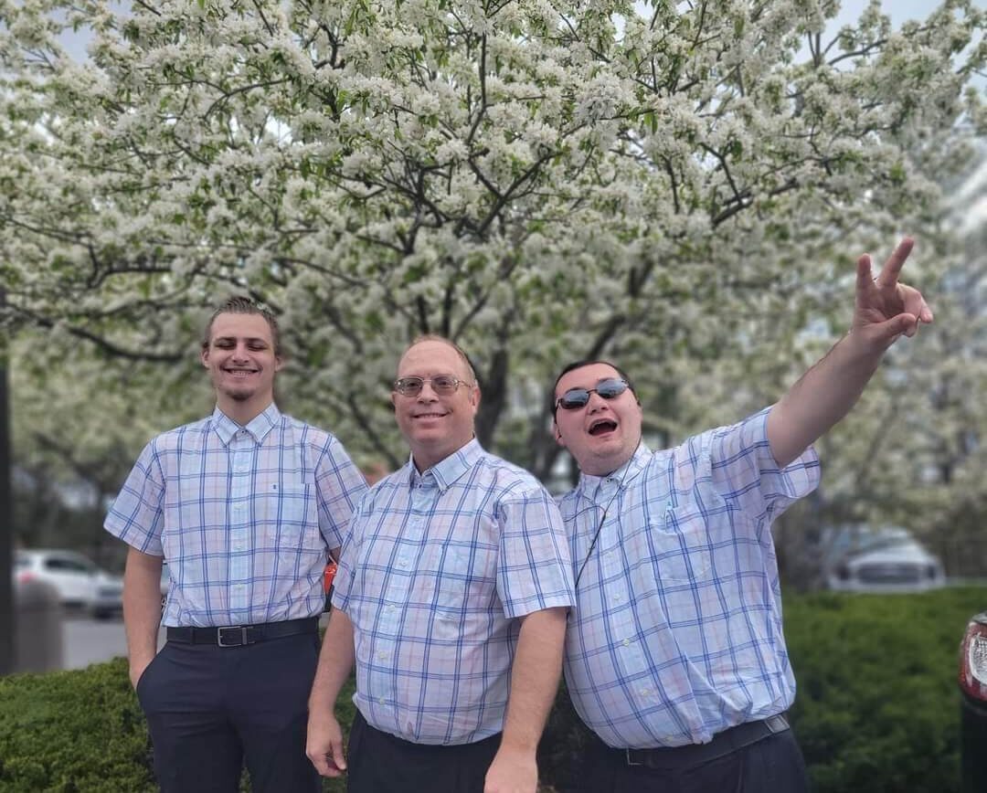 Three males in the same plaid shirt standing in front of a flowering tree, one pointing